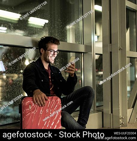 Smiling young man with suitcase sitting at rainy window using smartphone