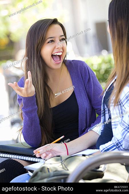 Attractive expressive young mixed-race female student sitting and talking with girlfriend outside on bench