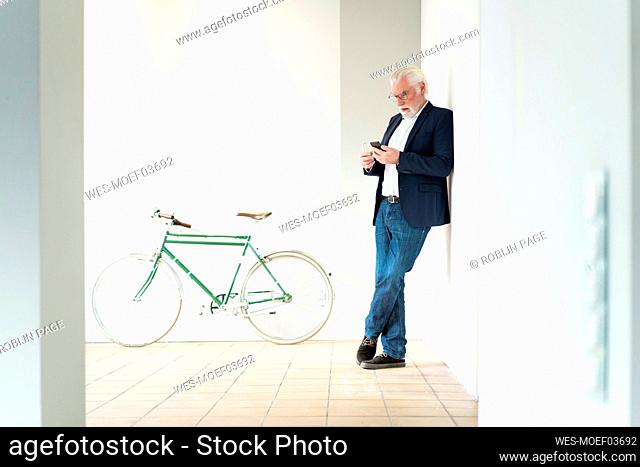Senior male entrepreneur using smart phone while leaning on wall at office