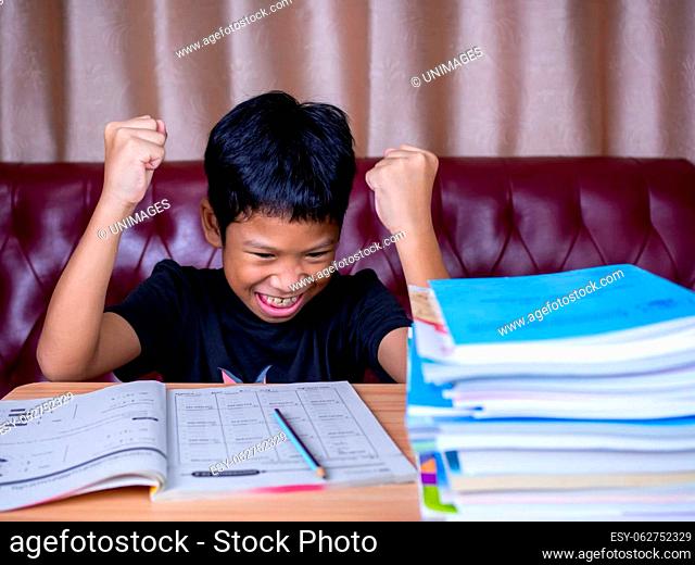 The boy was very happy to finish his homework. He sat on a wooden table and had a stack of books next to it. The background is a red sofa and cream curtains