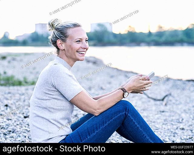 Happy mature woman holding mobile phone while sitting at riverbank