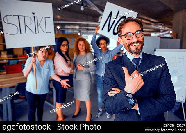 Laughing director showing thumb up, striking employees on backdrop. Modren loft office unsatisfied workers striking for their rights. Toned concept