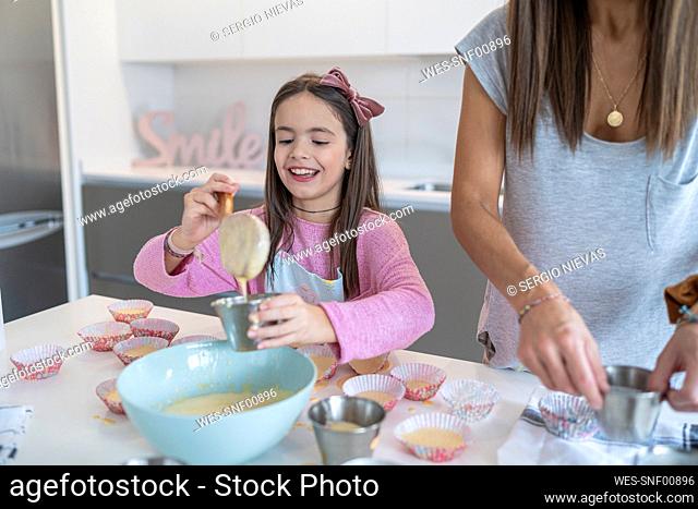 Smiling daughter scooping batter in cupcake holder by mother in kitchen