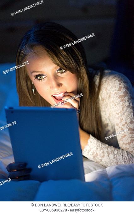 Teenage girl in the dark lying on a bed working on a tablet computer