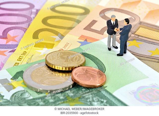 Miniature figurines of managers shaking hands while standing on euro banknotes and coins, symbolic image for business, sealing a contract