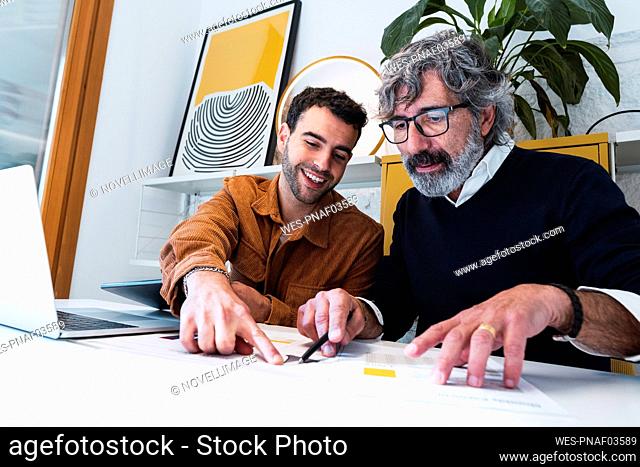 Smiling young freelancer explaining document to colleague sitting at table