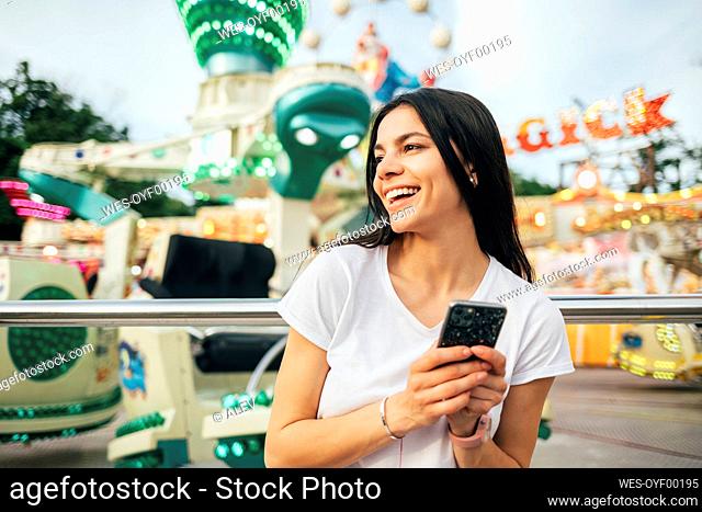 Cheerful young woman holding smart phone looking away at amusement park