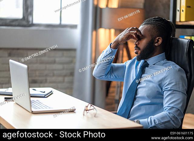 Tired young office worker seated at the table with his eyes closed touching his nose bridge