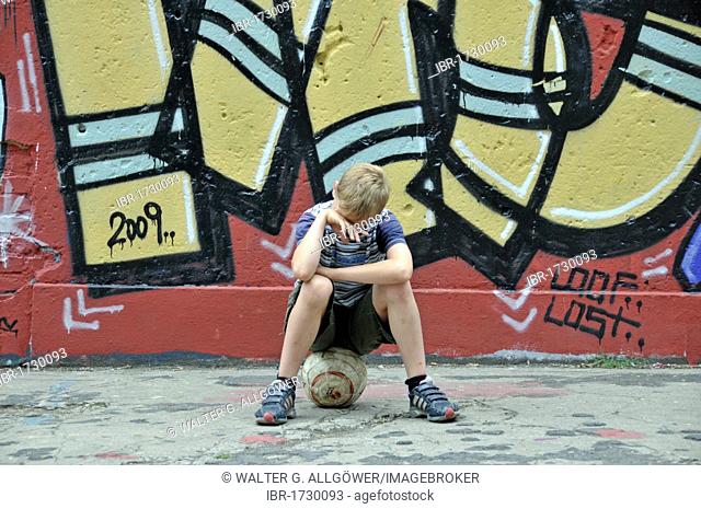 Disappointed and lonely boy with his football in front of a wall with graffiti, playground for football in Cologne, North Rhine-Westphalia, Germany, Europe