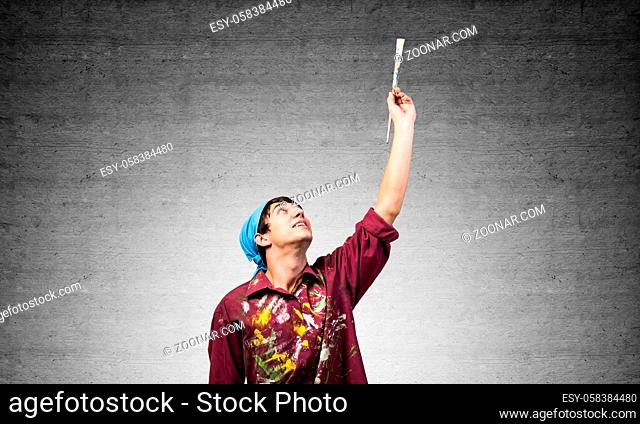 Young painter artist pointing upwards. Man holds paintbrush over his head. Portrait of caucasian painter on gray wall background