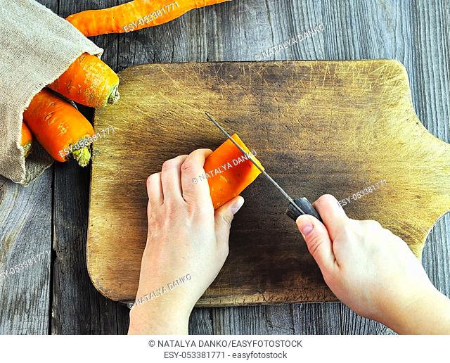Two female hands with a knife and carrot on a wooden kitchen board, top view