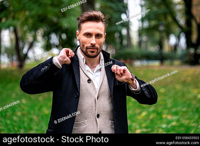 A handsome young man straightens the sides of his dark blue coat. A young business man stands on the street in an autumn coat and cardigan under it looks...