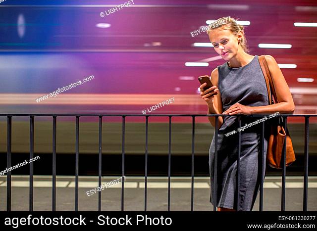 Pretty, young female commuter waiting for her daily train in a modern trainstation, using her cellphone while waiting (color toned image)