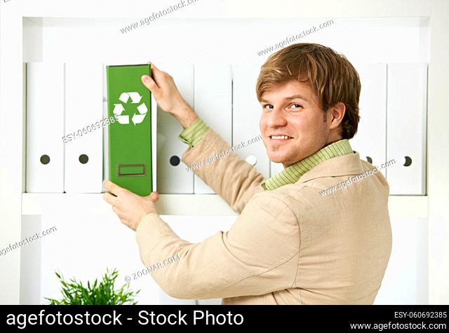 Businessman removing green folder with recycling symbol from shelf, looking back at camera, smiling