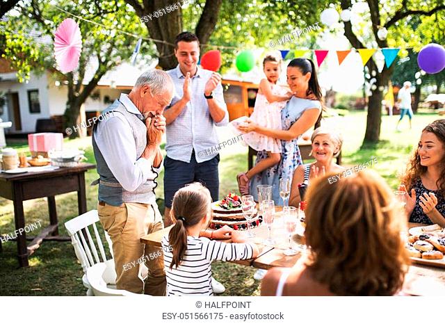 A senior man with an extended family looking at the birthday cake, crying in happiness. Celebration outside in the backyard or big garden party