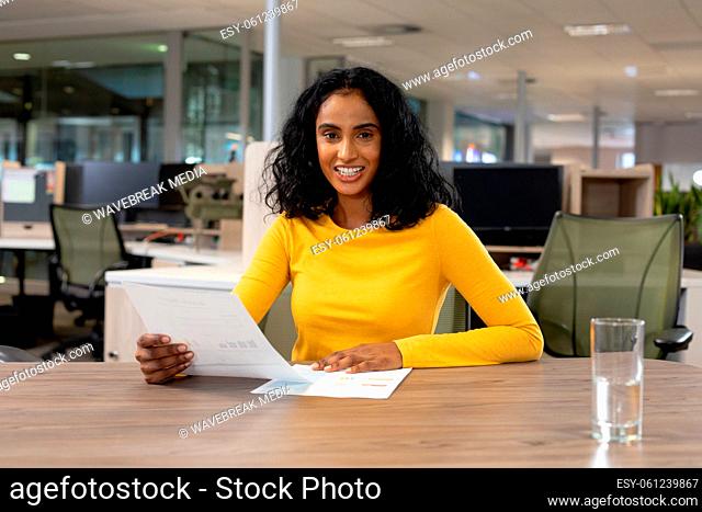 Portrait of smiling multiracial businesswoman with document sitting at desk working in office