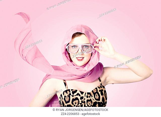 Beautiful retro fashion shot of a girl in late 20s in studio wearing a sleeveless leopard print top with pink headscarf blowing in the wind and 50s style shades