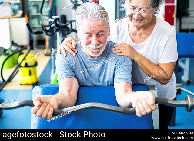 cute couple of two seniros at the gym doing exercises - man holding a barr with weight witgh his wife helping him and looking it