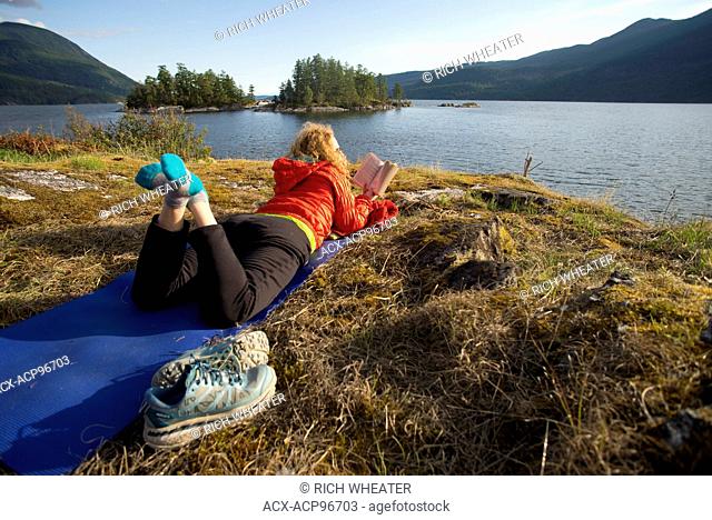 Woman reading book on yoga mat while camping at Kunechin Point. Sechelt Inlet. Sechelt, British Columbia. Canada