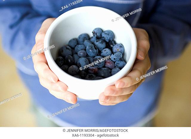 A boy holds a small white bowl containing freshly picked blueberries