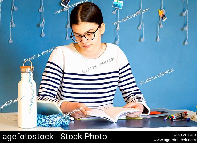 Portrait of girl sitting at desk with school book looking at digital tablet