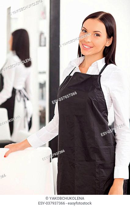 Attractive hairdresser is standing near a chair and mirror. She is inviting customers in her beauty shop. The woman is looking at the camera and smiling