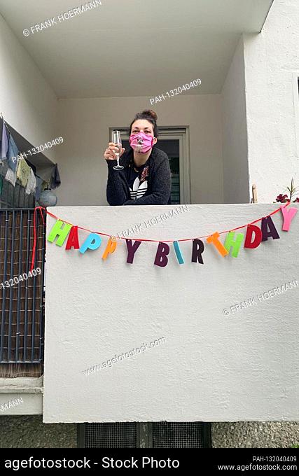 Lonely birthday in times of the coronavirus pandemic on April 28, 2020. A young woman stands alone on her small balcony on her birthday with a mask and holds a...