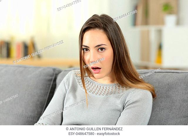 Portrait of a perplexed woman looking at camera sitting on a couch in the living room at home. She does not believe you