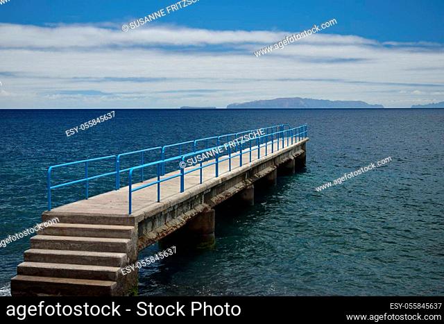 A little pier with stairs and a blue handrail at the ocean. Santa Cruz, Madeira, Portugal. Islands in the background