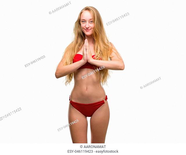 Blonde teenager woman wearing red bikini praying with hands together asking for forgiveness smiling confident