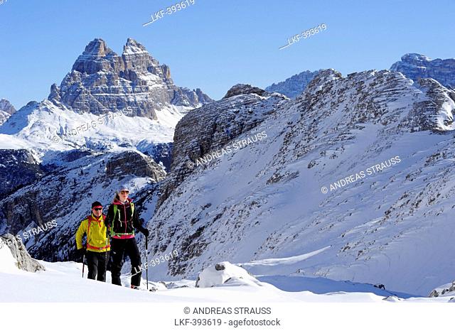 Young woman and young man ascending with crosscountry skis to Corno d'Angolo, Tre Cime di Lavaredo in background, Corno d'Angolo, Cortina, Veneto, Dolomites