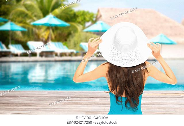 people, summer holidays, travel, tourism and vacation concept - woman in swimsuit and sun hat from back over exotic hotel resort beach with swimming pool and...