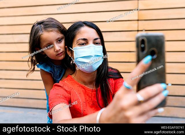 Mother wearing mask taking selfie with daughter against wall