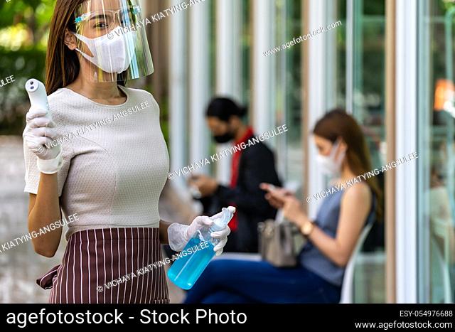 Portrait attractive asian waitress wear face mask and shield hold thermometer and alcohol gel sanitizer with background of social distancing customer waiting in...