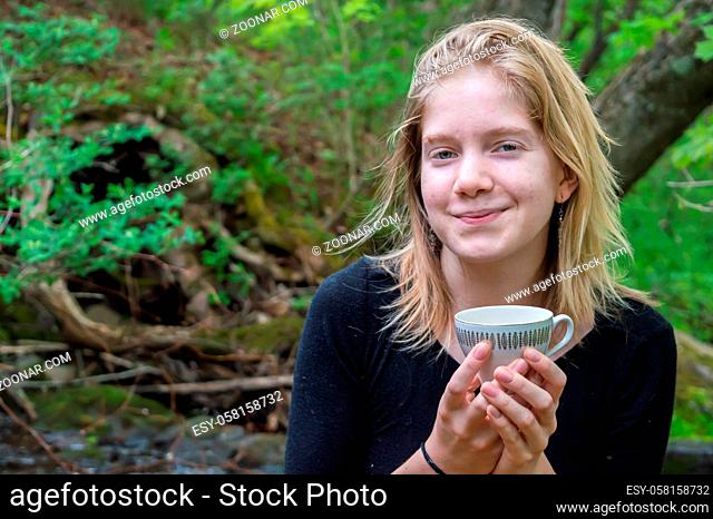 Portrait of a pretty young girl sitting on a rock on the shore of a forest river, holding a china tea cup with both hands and smiling at camera