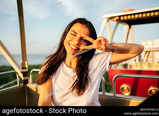 Smiling beautiful woman showing peace sign and winking while enjoying Ferris wheel ride