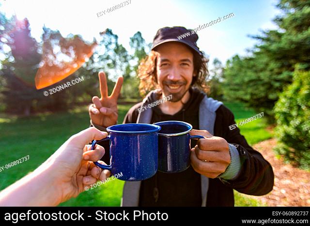 Selective focus of two happy persons clinching their cups of coffee and tea together while standing in a national park full of nature scenery