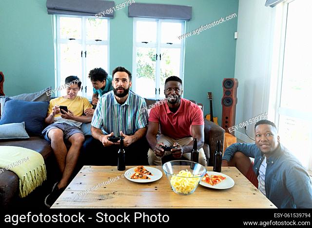 Multiracial male friends with beer bottles and food on table playing video game on sofa at home