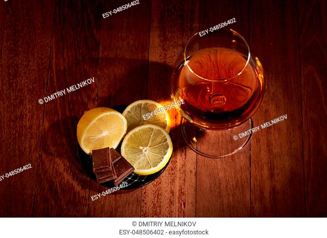 Still life with glass with cognac and plate with pieces of lemon and chocolate standing on a table on a dark wooden background. Top view