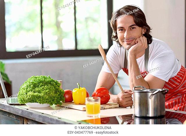 Joyful young man is waiting for prepared food in kitchen. He is standing and holding wooden spoon near pot. Chef is looking at camera and smiling