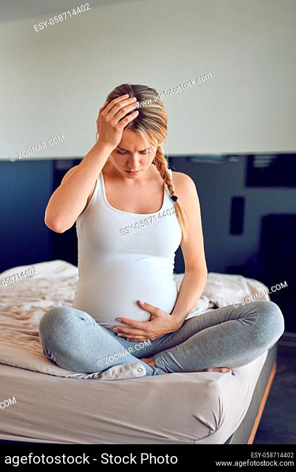 Troubled heavily pregnant young woman sitting cross legged on a bed cradling her swollen abdomen and clutching her head looking down with a serious expression