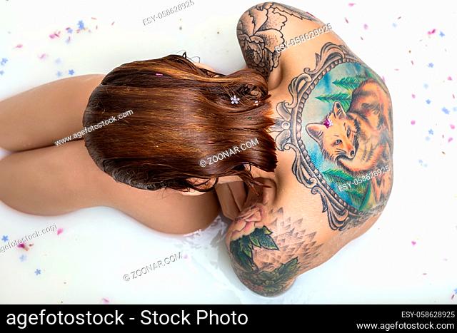 Redhead girl with wet hair sits in the bath full of water with milk and flower petals. She has a colorful fox tattoo on the back and another tattoos on her...