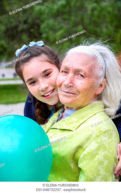Beautiful girl hugging his beautiful gray-haired grandmother. A pleasant elderly woman sits on a park bench holding a balloon and they both smile happily