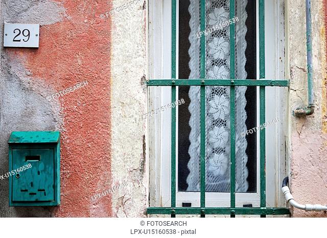 Closeup view of heavily textured pink wall, green letter box, and green metal window railings with white lace hangin behind frosted glass, Monterosso al Mare
