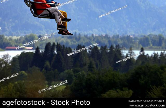 13 June 2022, Bavaria, Buching: Excursionists ride in the sunshine in a chairlift on the Buchenberg in front of Forggensee