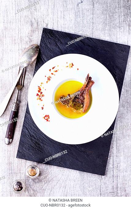 White ceramic plate with well done grilled lamb chop in yellow broth with chili pepper's flackes over black slate board over white wooden table