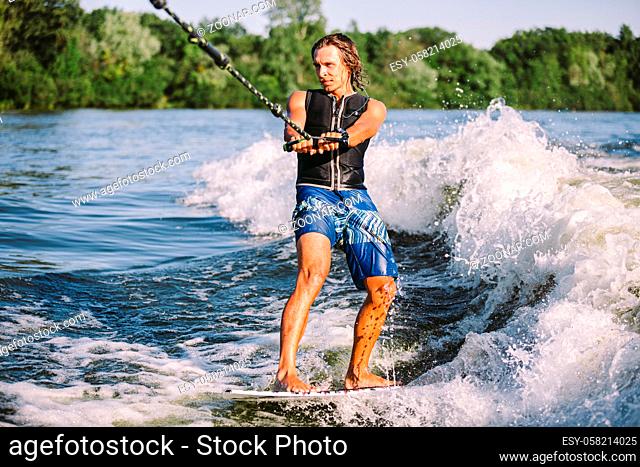 Young athletic man with long hair wakesurfing on waves of river in sunny summer weather. Ttheme outdoor activities in summer