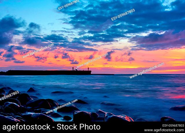 Seascape with Atlantic ocean and the sky at twilight, stones on the beach and fishermans on the pier. Long exposition with blurred sea water