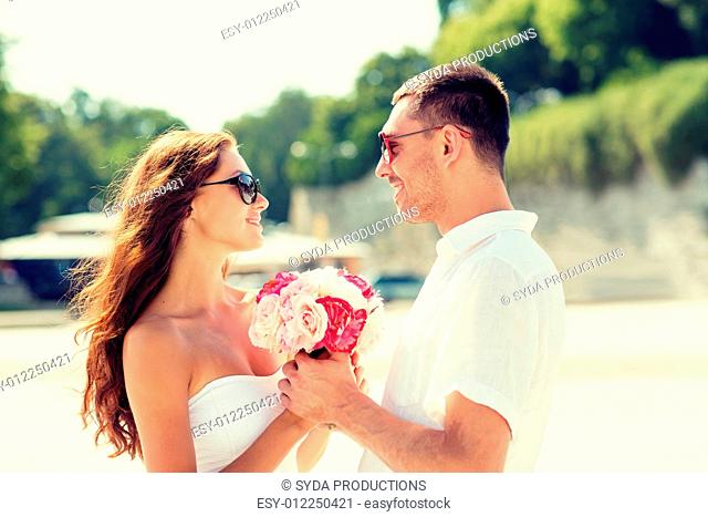 love, wedding, summer, dating and people concept - smiling couple wearing sunglasses with bunch of flowers looking at each other in city