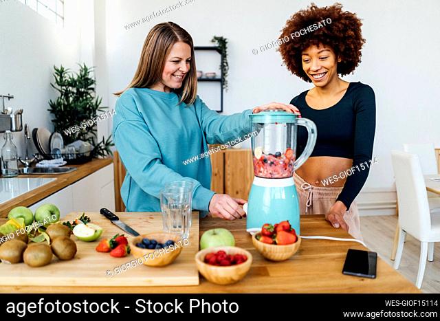 Happy young woman standing by friend preparing smoothie in kitchen